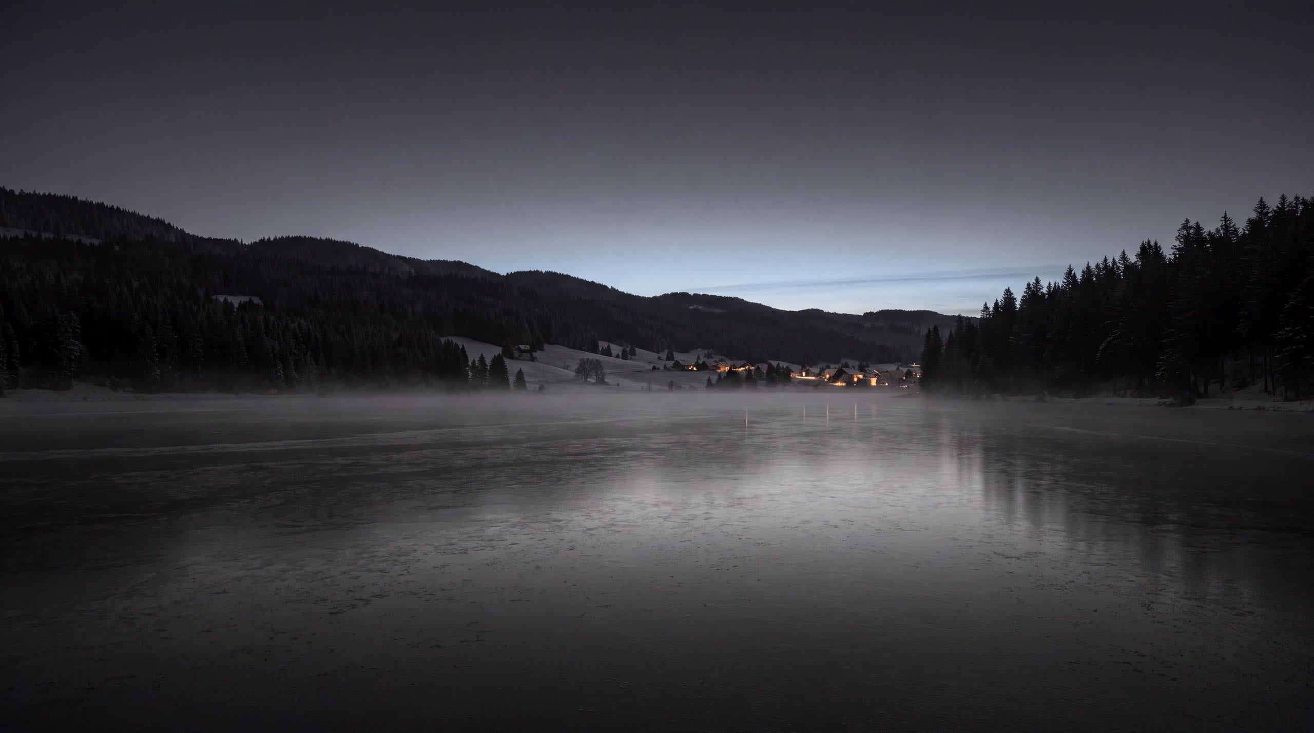 The frozen Lac de Joux at dawn: mist on the ice, forested hills on both shores and the lights of a village at the far end of the valley.