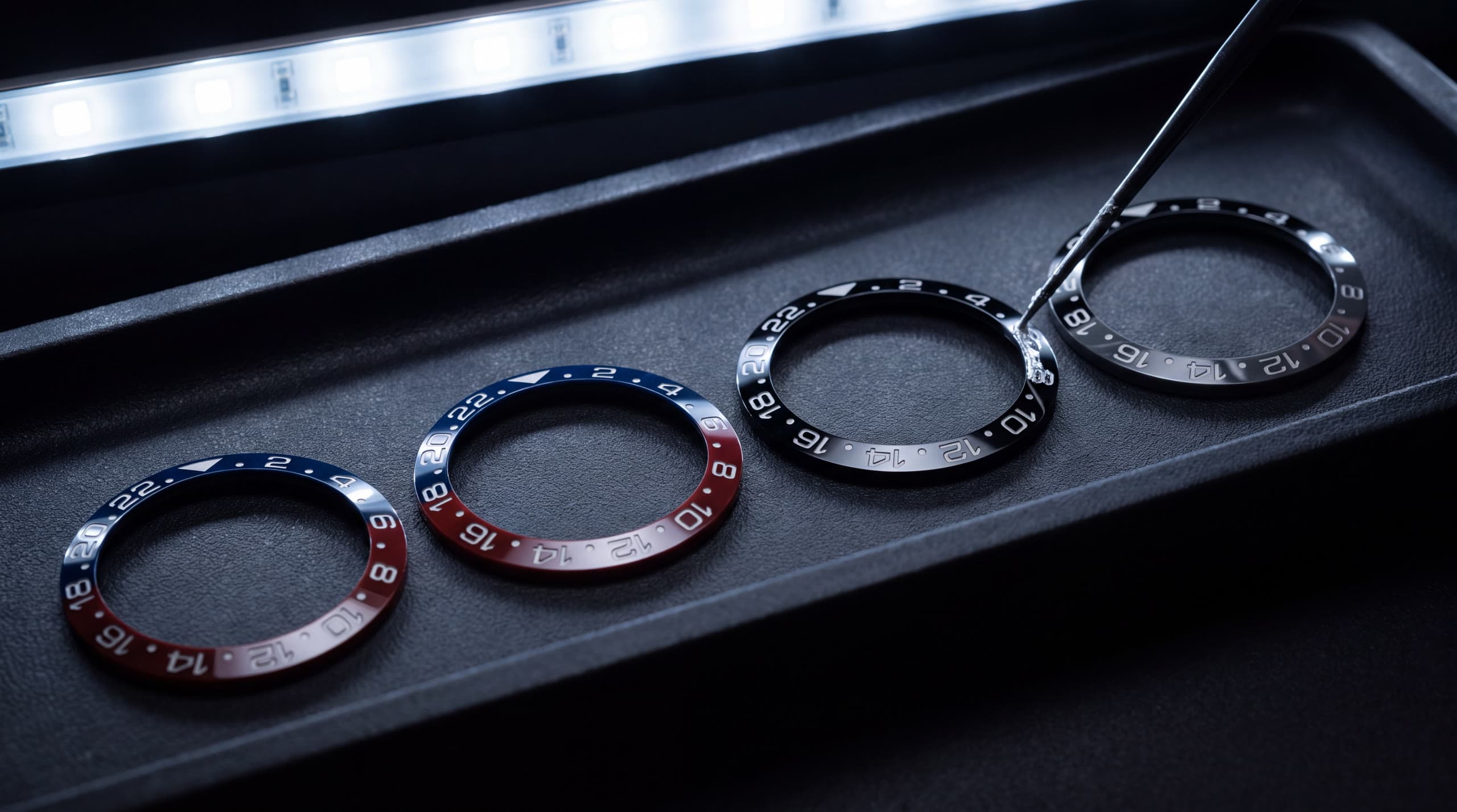 Four engraved ceramic bezel inserts laid out on a black finishing tray under a strip light