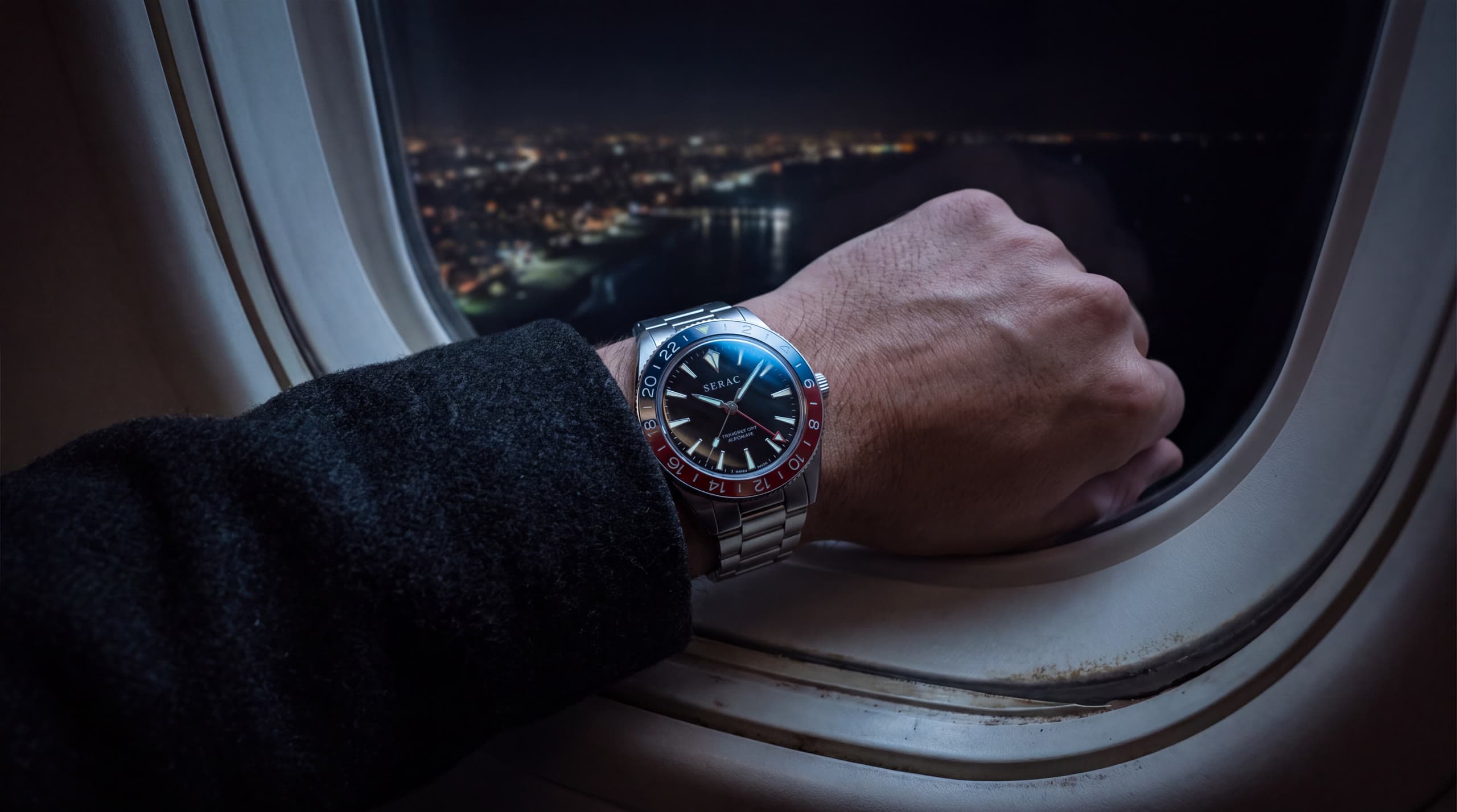 A wrist resting at an aircraft window at night, city lights far below; the Traverse GMT with its blue and red bezel.