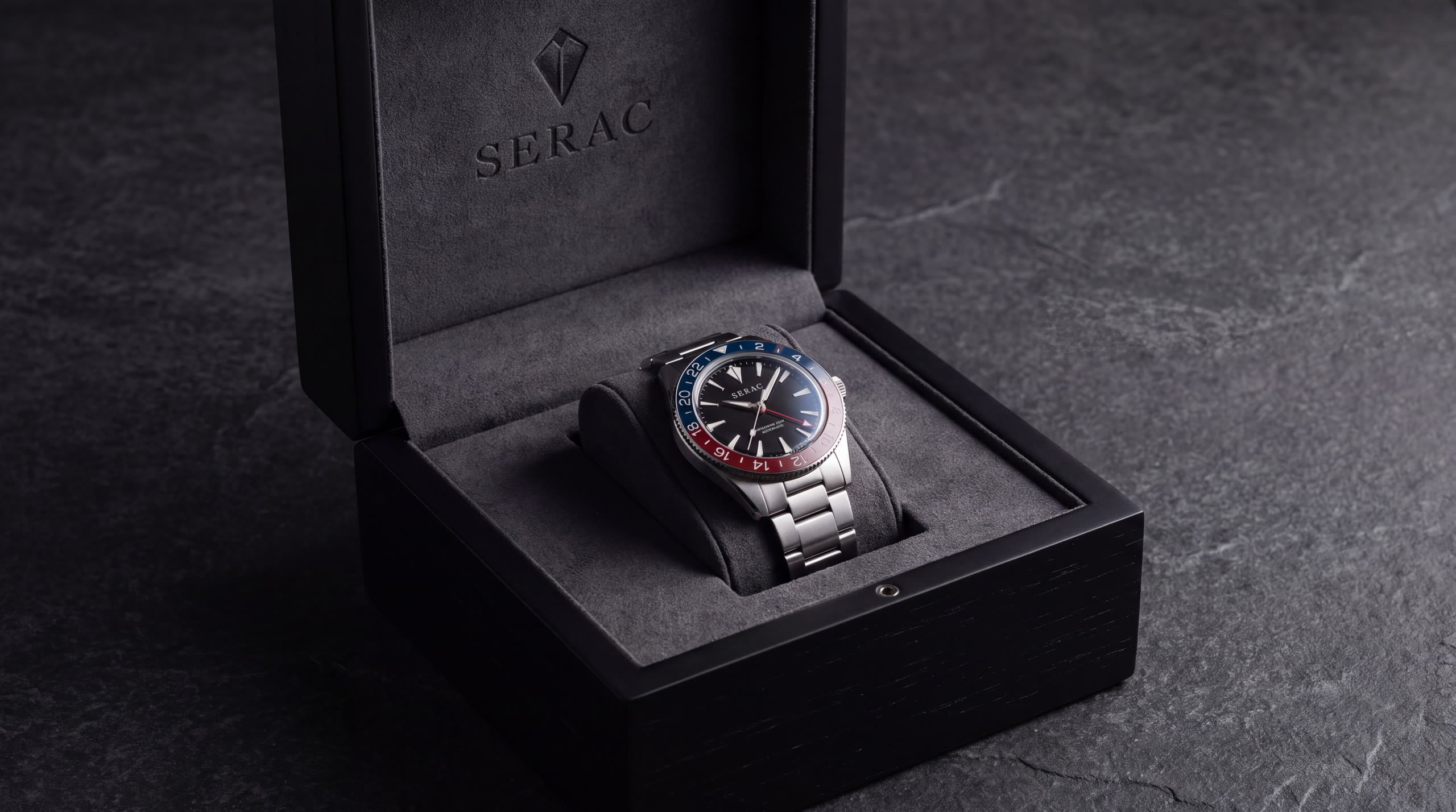 A Traverse GMT with a blue and red bezel resting on a grey cushion inside an open black presentation case embossed with the SERAC peak, on dark slate.