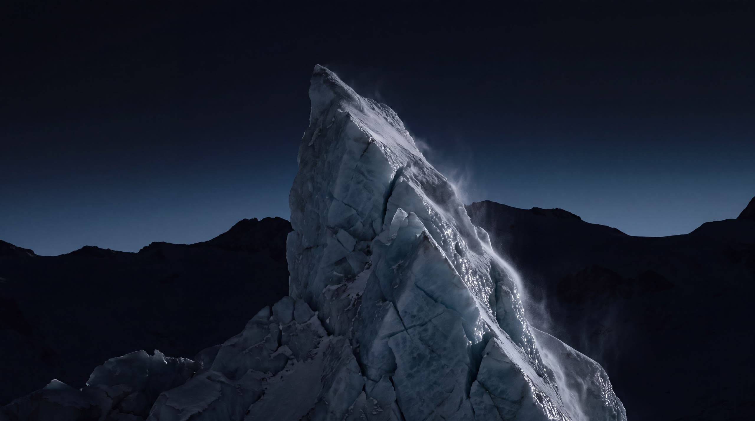 A tower of glacial ice at blue hour, wind lifting snow from its crest against dark mountains.
